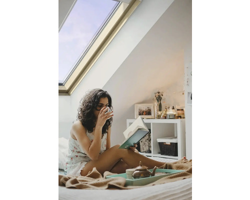 Žena čte knihu v posteli pod střešním oknem.