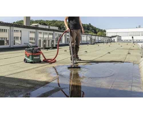 Muž vysává vodu z ploché střechy pomocí mokrého a suchého vysavače. V pozadí je budova.