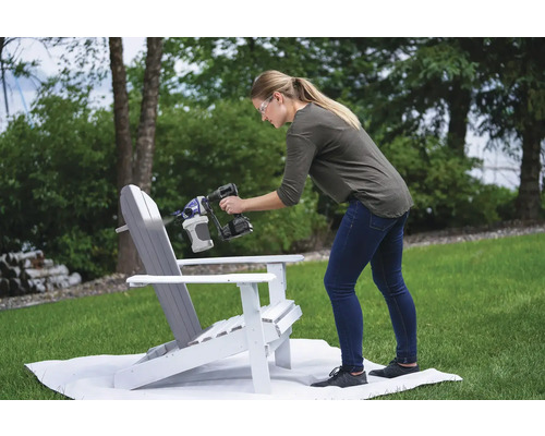 Žena natírá dřevěnou židli stříkací pistolí na barvu na zahradě