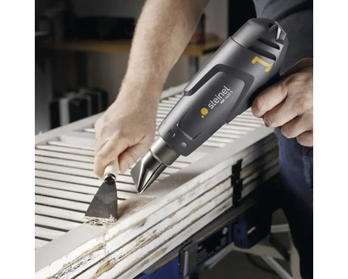 Řemeslník odstraňuje barvu z dřevěných dveří horkovzdušnou pistolí a špachtlí