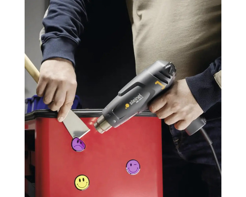 Osoba odstraňuje nálepky z povrchu pomocí horkovzdušné pistole a špachtle.