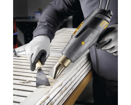 Osoba odstraňuje barvu z dveří pomocí horkovzdušné pistole a špachtle.