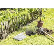 Zahradní scéna s dřevěným plotem, mladým stromem v květináči, rýčem a drátěným pletivem na ochranu rostlin.