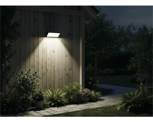 Venkovní pohled na dům se svítící nástěnnou lampou, obklopený rostlinami a chodníkem