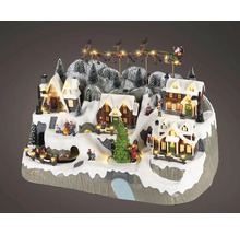 Osvětlená vánoční vesnice se zasněženými domy, jedlemi, figurkami, vlakem a letícími sáněmi Santa Clause.