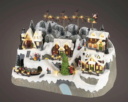 Osvětlená vánoční vesnice se zasněženými domy, jedlemi, figurkami, vlakem a letícími sáněmi Santa Clause.
