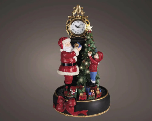 Vánoční dekorace s postavičkou Santa Clause a dítěte zdobícího stromeček u zdobených hodin na kulatém podstavci s červenou mašlí.