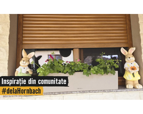 Truhlík s muškáty a dvěma figurkami zajíců na okenním parapetu před dřevěnou roletou.