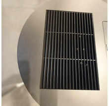 Detailní záběr na obdélníkovou černou kovovou ventilační mřížku integrovanou do kruhového povrchu z nerezové oceli.