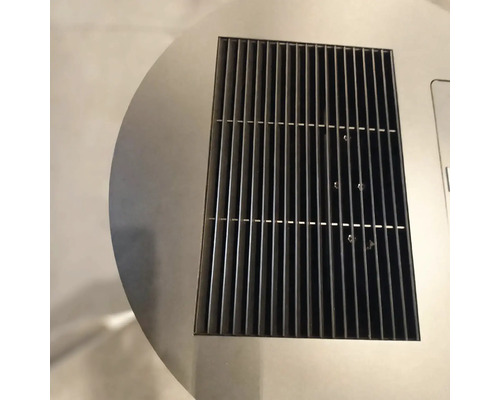 Detailní záběr na obdélníkovou černou kovovou ventilační mřížku integrovanou do kruhového povrchu z nerezové oceli.