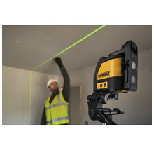Stavební dělník v pracovním oděvu používá křížový laser DeWalt na stativu k přesnému zaměření zelené laserové linie na stěně.