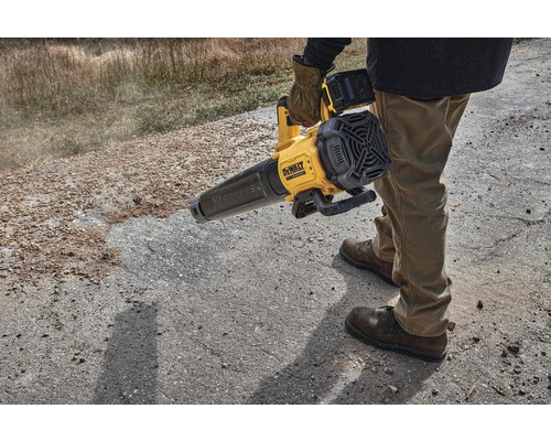 Osoba obsluhuje žlutý akumulátorový foukač listí DeWalt na nezpevněném povrchu.