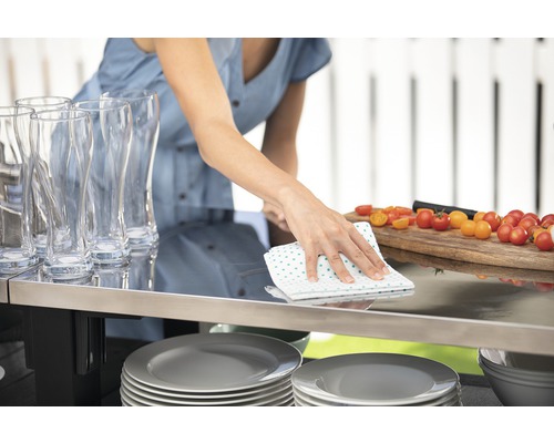 Žena utírá stůl utěrkou, nádobí, sklenice a rajčata na obrázku.