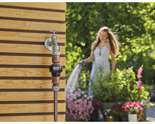 Měřič zahradní vody na vodovodním kohoutku se ženou v pozadí, která zalévá zahradu