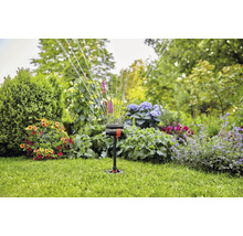 Zahradní postřikovač v provozu na trávníku s květinovými záhony v pozadí.