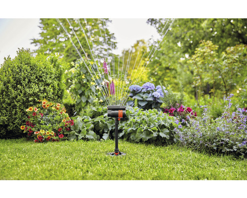 Zahradní postřikovač v provozu na trávníku s květinovými záhony v pozadí.