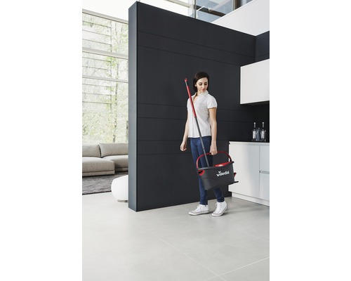 Žena nese kbelík a mop v moderním obývacím pokoji.