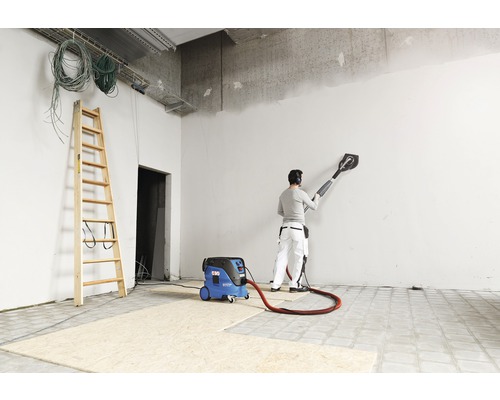 Muž brousí stěnu pomocí brusky s dlouhým krkem a vysavače v místnosti se žebříkem.