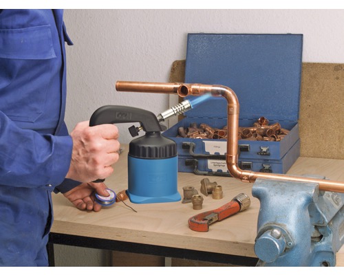 Muž pájí měděné trubky pájecí lampou, armaturami a nářadím na ponku