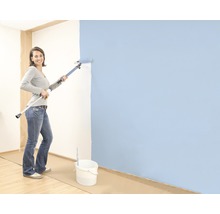 Žena maluje stěnu válečkem na teleskopické tyči, na podlaze stojí kbelík s barvou.