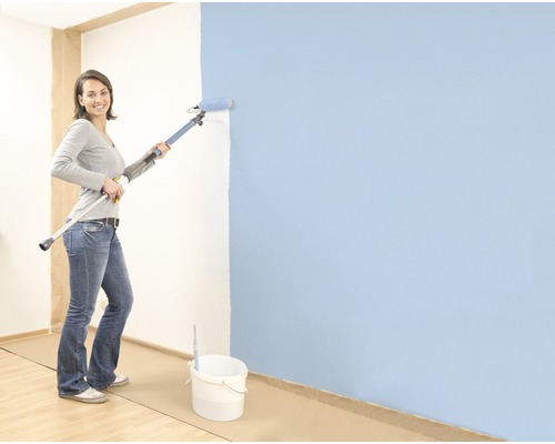 Žena maluje stěnu válečkem na teleskopické tyči, na podlaze stojí kbelík s barvou.