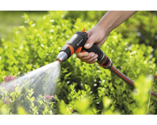 Ruka drží zahradní stříkací pistoli značky Gardena, ze které stříká voda na rostliny.