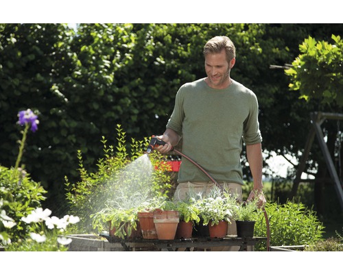 Muž zalévá rostliny na zahradě zahradní stříkačkou a zahradní hadicí.