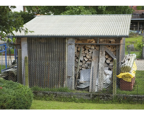 Dřevěná kůlna s palivovým dřívím a vlnitou plechovou střechou za pletivovým plotem