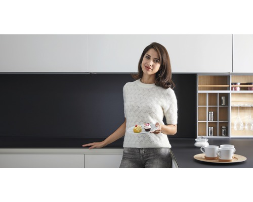 Moderní kuchyně s bílými skříňkami a tmavou pracovní deskou, žena s talířem a pečivem