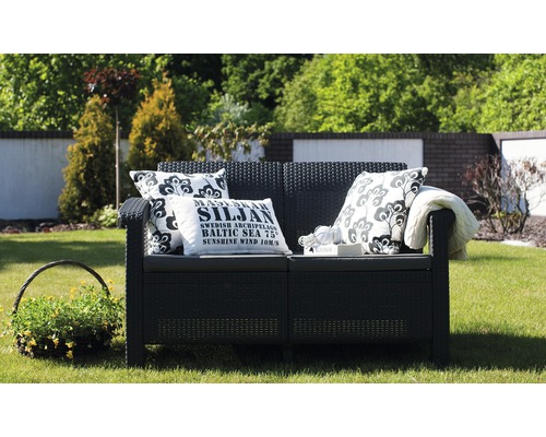 Zahradní scéna s černou zahradní lavicí, dekorativními polštáři a košíkem na rostliny