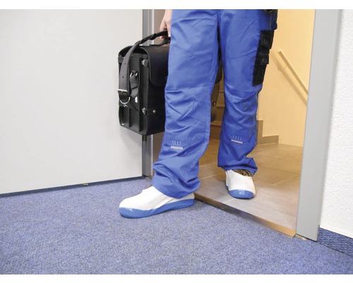 Osoba v modrých pracovních kalhotách, návlecích na boty a s kufříkem na nářadí vstupuje do místnosti.