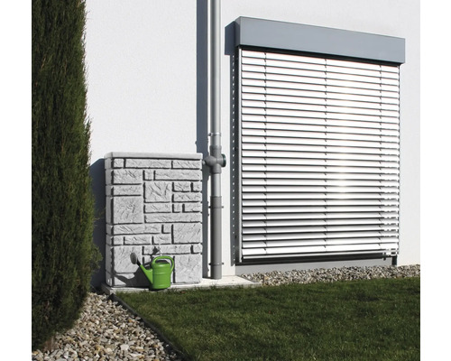 Nádrž na dešťovou vodu s kamenným vzorem a zelená konev vedle svodu před stěnou domu