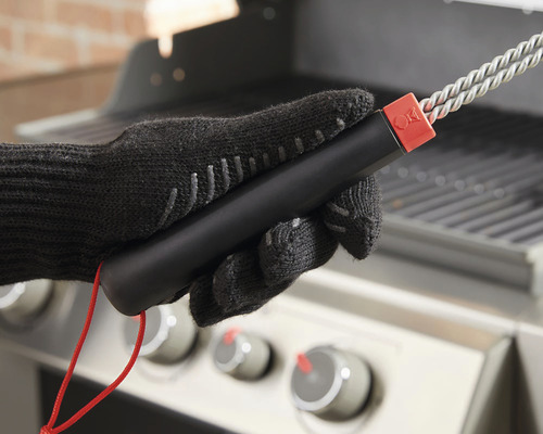 Ruka v grilovací rukavici drží grilovací kartáč před grilem.