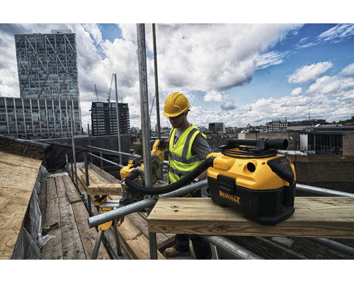 Řemeslník s ochrannou helmou a výstražnou vestou pracuje s mokro-suchým vysavačem Dewalt na lešení před městskou scenérií.