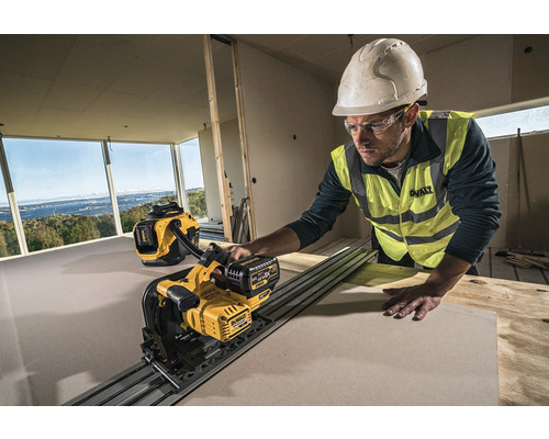 Stavební dělník s helmou a ochrannými brýlemi řeže desku sádrokartonovou pilou DeWalt v hrubém prostoru. V pozadí je vysavač DeWalt.