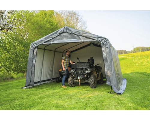 Šedý přístřešek na nářadí s mužem a čtyřkolkou na louce.