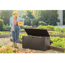 Žena s dřevěnou bednou a zahradní lopatkou stojí vedle otevřeného zahradního boxu na zahradě.