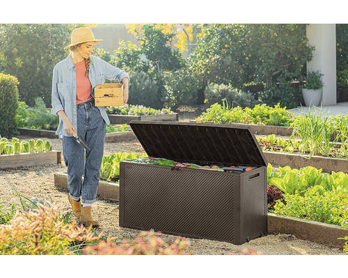 Žena s dřevěnou bednou a zahradní lopatkou stojí vedle otevřeného zahradního boxu na zahradě.