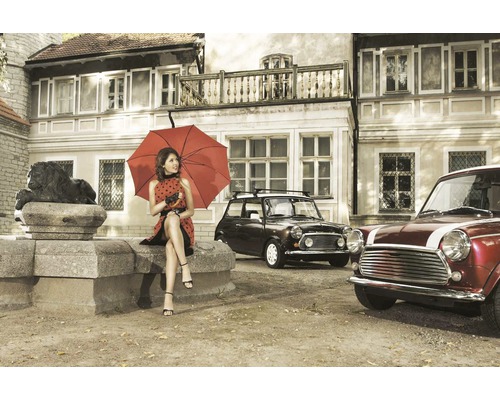 Žena s deštníkem sedí před budovou se starými auty.