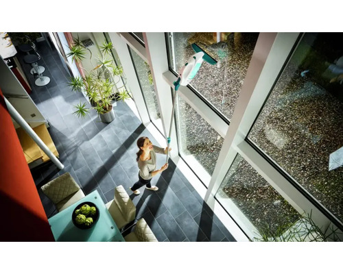 Žena myje okna čističem oken s teleskopickou tyčí v prosvětleném obývacím pokoji.