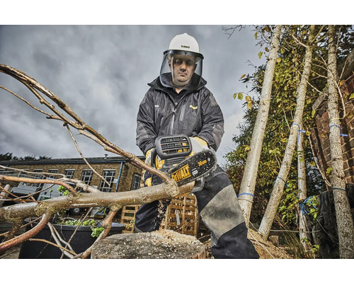 Osoba v ochranném oděvu a přilbě řeže větev akumulátorovou pilou. V pozadí jsou vidět stromy.