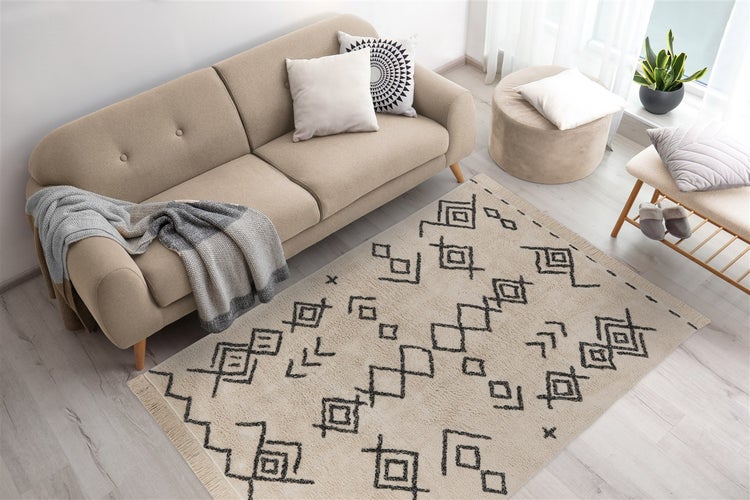 Obývací pokoj s béžovou pohovkou, pufem a kobercem s černými geometrickými vzory a třásněmi na světlé podlaze.