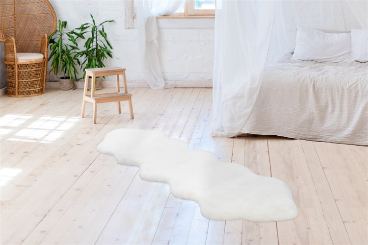 Bílý běhoun ve vzhledu ovčí kůže na světlé dřevěné podlaze v prosvětlené ložnici s postelí a proutěným křeslem.