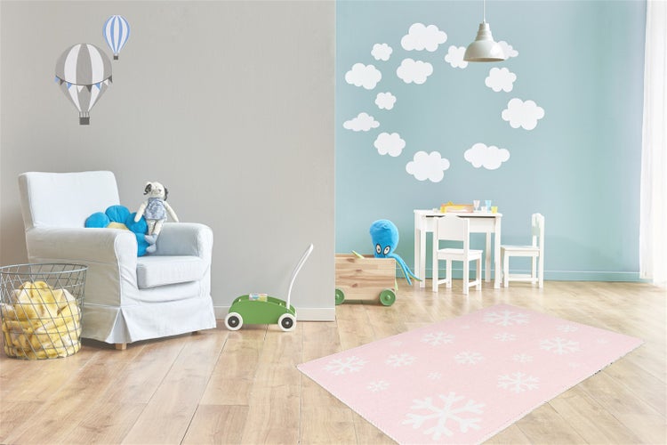 Světlý dětský pokoj s růžovým kobercem se vzorem sněhových vloček na dřevěné podlaze, křeslem, dětským stolkem se židlemi a samolepkami na zeď.