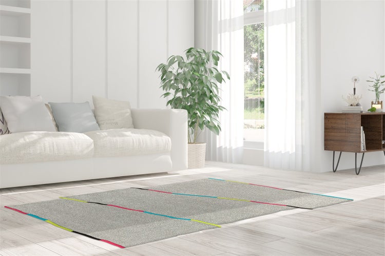 Světlý obývací pokoj s šedým kobercem s barevnými diagonálními liniemi na světlé dřevěné podlaze, bílou pohovkou a rostlinou.