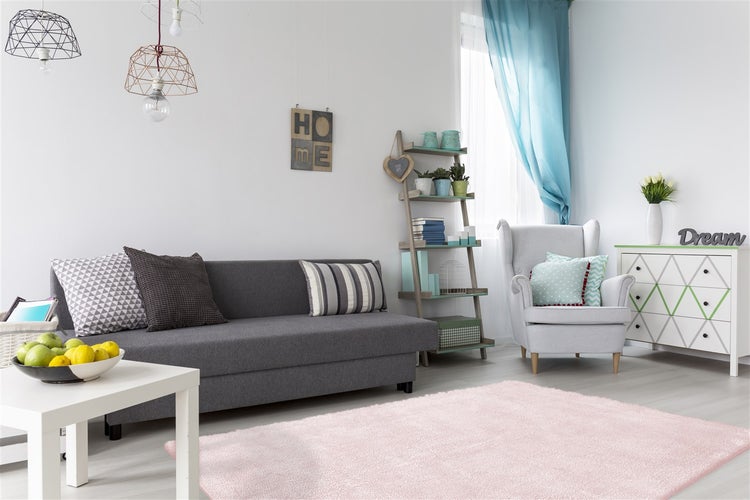Světlý obývací pokoj s šedou pohovkou, růžovým kobercem s vysokým vlasem, světlým ušákem a komodou. Moderní závěsná svítidla a policový žebřík.