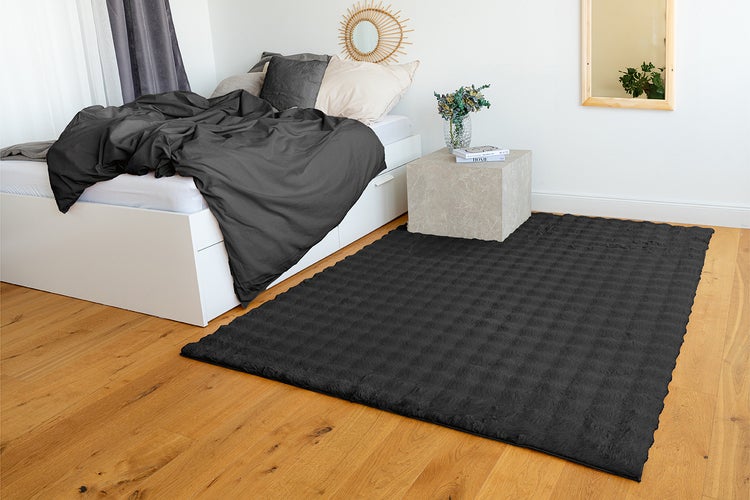Moderní antracitový strukturovaný koberec na dřevěné podlaze ve světlé, organizované ložnici s bílou postelí a kamenným nočním stolkem.