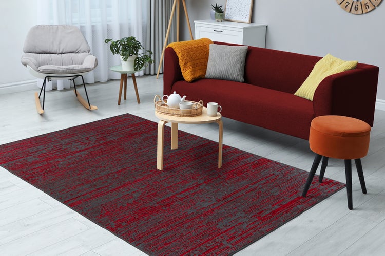 Moderní obývací pokoj s červeno-šedým vzorovaným kobercem na světlé podlaze, červenou pohovkou, šedým houpacím křeslem a dřevěným stolkem.