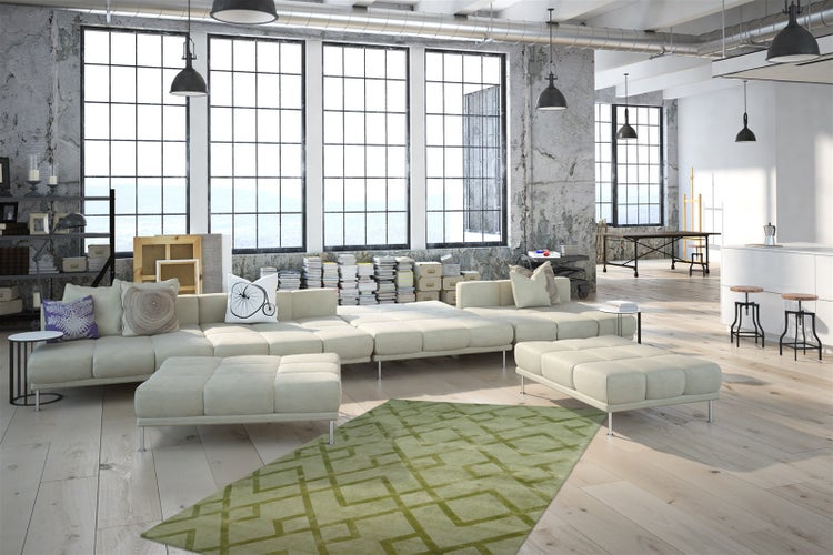 Prostorný industriální loft s velkou modulární pohovkou, zeleným kobercem s geometrickým vzorem, dřevěnou podlahou a velkými okny u betonových zdí.