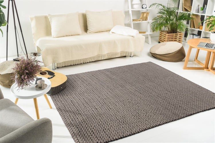Obdélníkový tmavě šedý tkaný koberec ve světlém, moderně zařízeném obývacím pokoji se světlou pohovkou, dřevěným nábytkem a rostlinami.